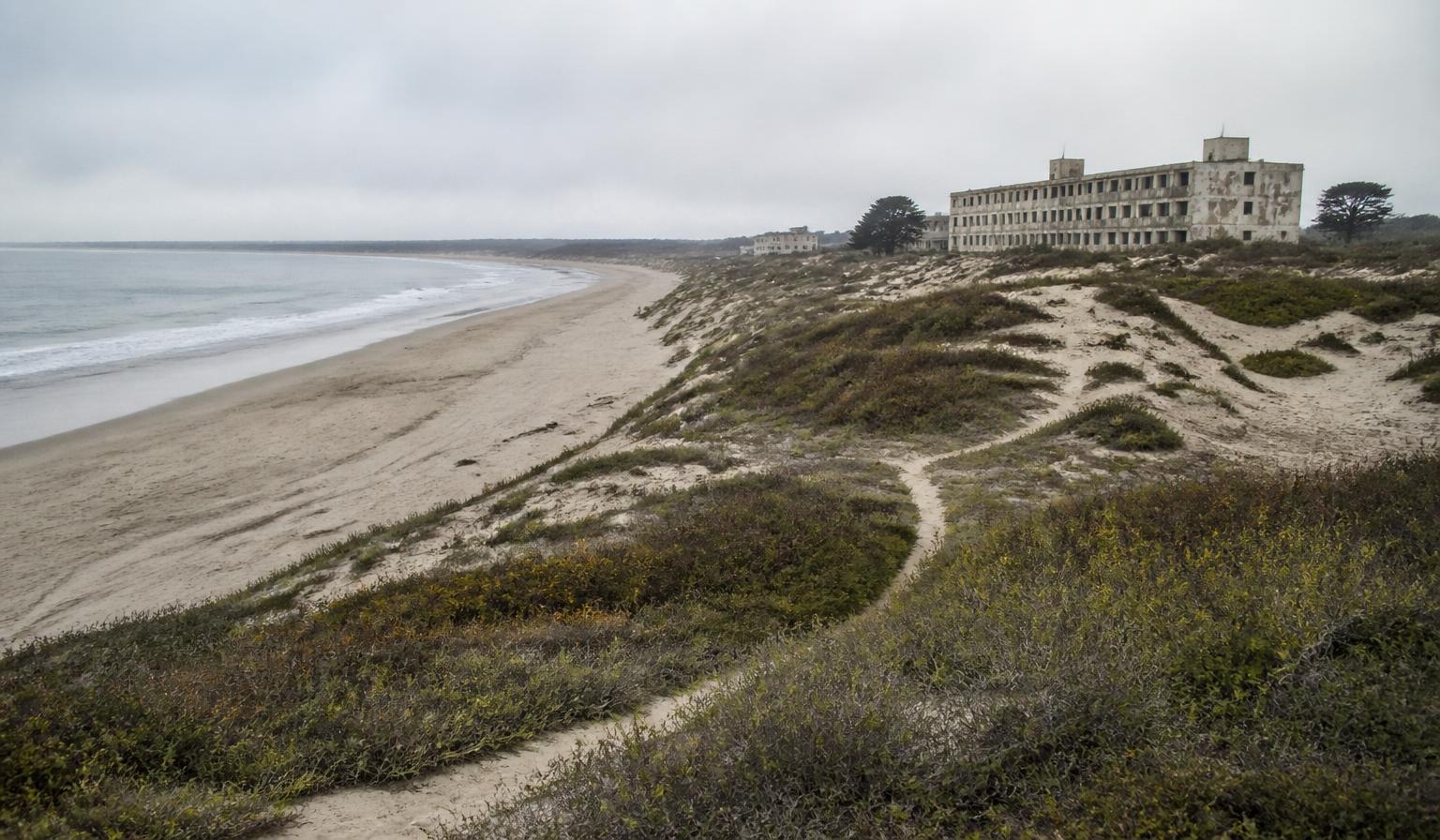 Aerial view of the former Fort Ord coastline near Monterey Bay, California, showing weathered barracks and coastal bluffs under overcast sky.